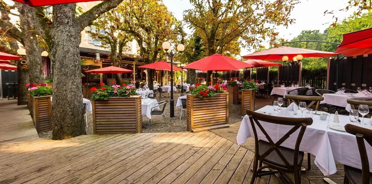 Gartenterrasse mit roten Sonnenschirmen unter alten Bäumen im warmen Nachmittagslicht.